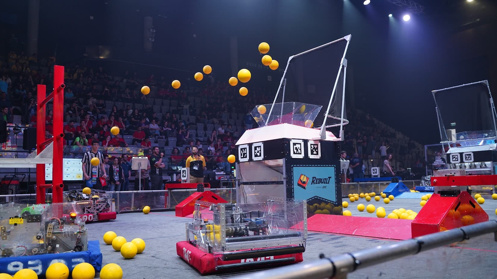 תמונה של 2 רובוטי FIRST Robotics Competition תופסים קוביות משחק שברקע קהל.