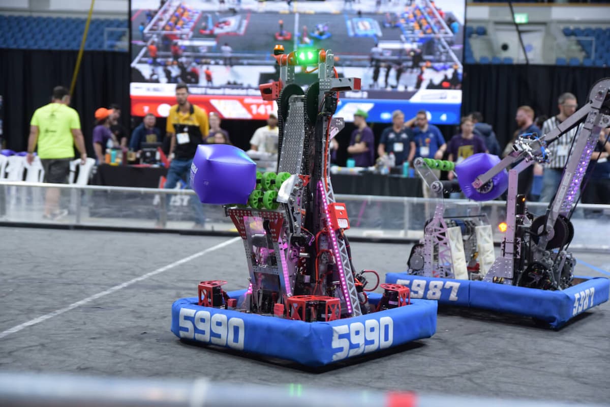 תמונה של 2 רובוטי FIRST Robotics Competition תופסים קוביות משחק שברקע קהל.